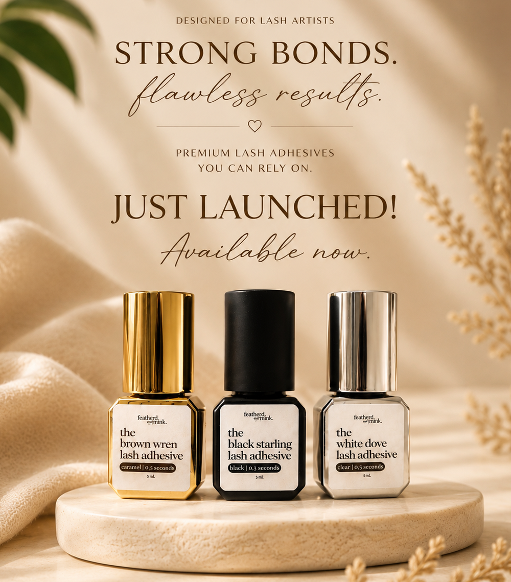 Three Feather Mink lash adhesive bottles displayed on a round stone platter with promotional text above. The bottles are labeled 'the brown wren lash adhesive,' 'the black starling lash adhesive,' and 'the white dove lash adhesive,' with gold, black, and silver caps against a warm beige background with soft fabric and dried wheat props.